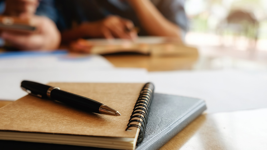 En una mesa, personas estudiando, libro, cuaderno y bolígrafo en primer término